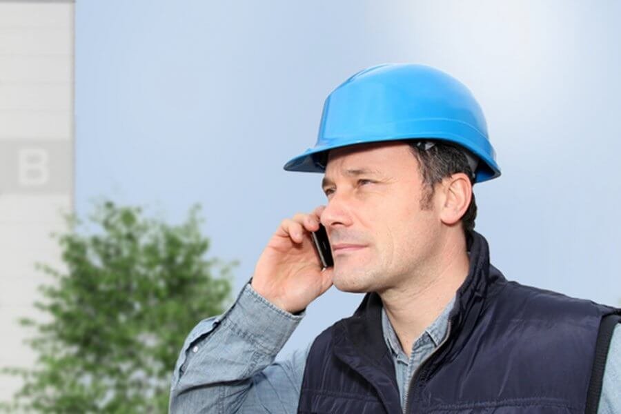 Ein Bauarbeiter mit hellblauem Sicherheitshelm steht vor einer weißen Wand und telefoniert.