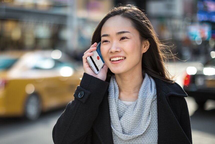 Eine Asiatin mit langen, braunen Haaren und einem grauen Strickpullover steht an einer stark befahrenen Straße und telefoniert.