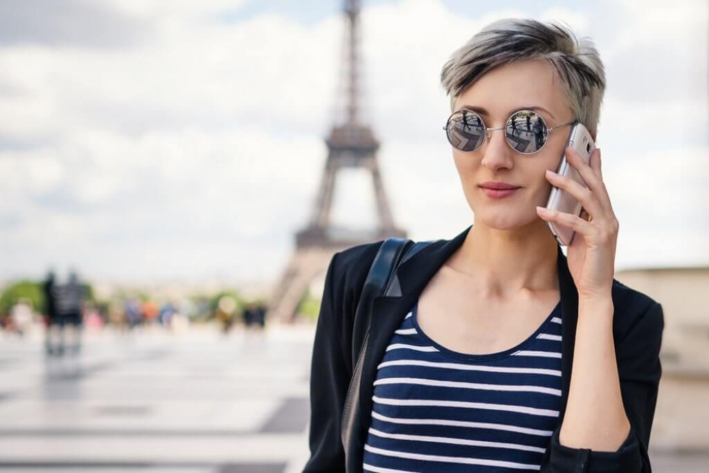 Eine junge Frau mit blonden kurzen Haaren, dunkelrotem Lippenstift und einer Sonnenbrille telefoniert mit dem Smartphone in Paris vor dem Eifelturm.