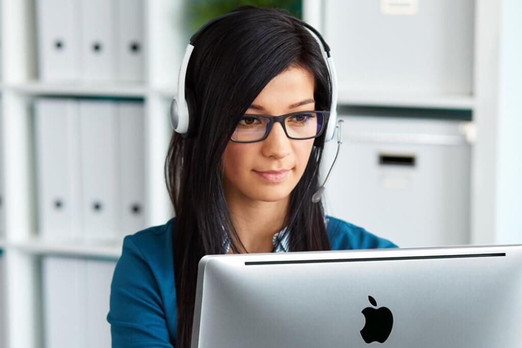 Eine Mitarbeiterin mit Sekretärinnenbrille und langen dunkelbraunen Haaren arbeitet am Laptop und trägt ein Headset.